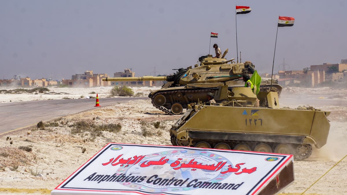 Egyptian tank and armoured personnel carrier flying Egyptian flags pass an Amphibious Control Command sign in the desert