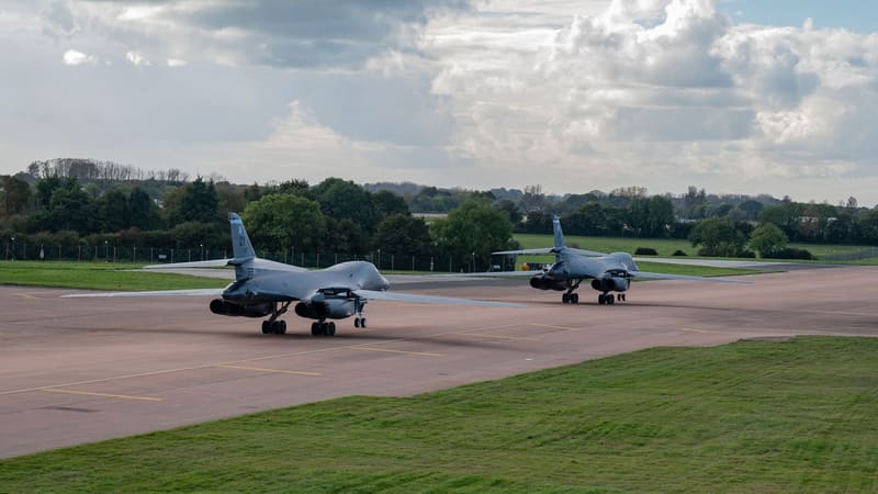 Two grey B-1B Lancer bombers taxi on an airfield taxiway past a perimeter fence, trees and fields under cloud