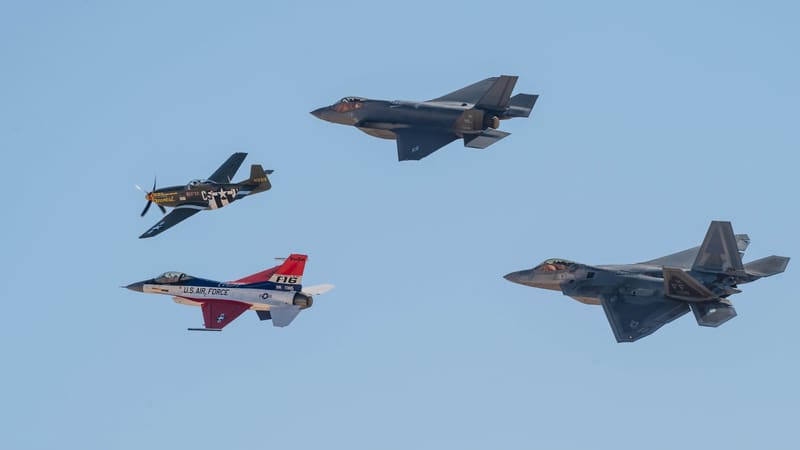 An F-35A, an F-22 and a red, white and blue F-16 fly in formation with a P-51 Mustang against a blue sky.