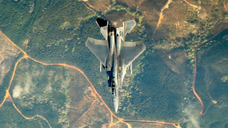 An F-15EX Eagle II seen from directly above, flying over green hills crossed by orange dirt roads.