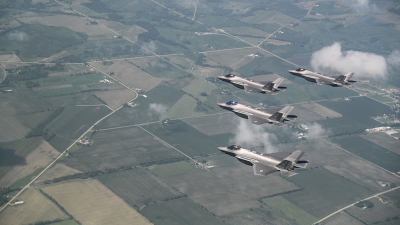 Four grey F-35A stealth fighters fly in close formation above green Wisconsin farmland.