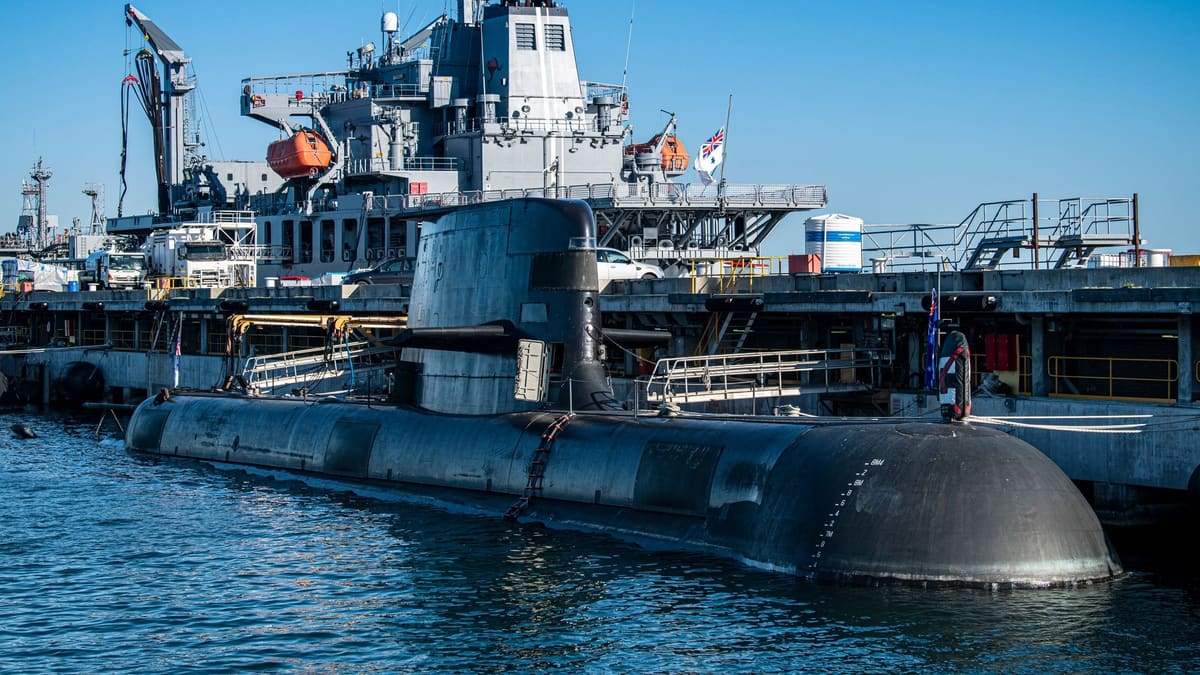 Dark grey Collins class submarine moored alongside a pier, with a supply ship and clear blue sky behind