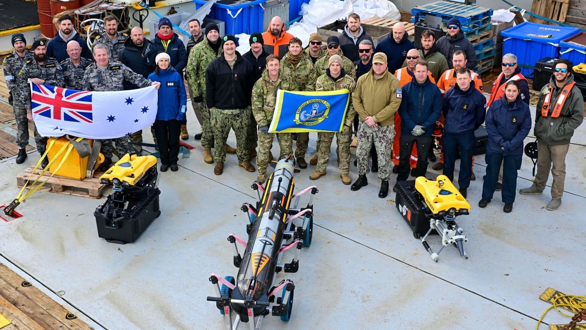 Navy personnel on a ship's deck hold an Australian naval ensign behind a torpedo-shaped underwater drone and two yellow ROVs