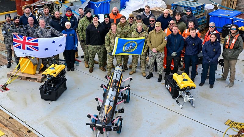Navy personnel on a ship's deck hold an Australian naval ensign behind a torpedo-shaped underwater drone and two yellow ROVs