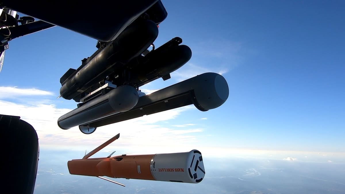 Orange Altius-700 drone test vehicle falling away from a launcher under a helicopter, with sky and ground below
