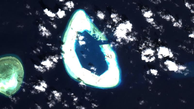 Satellite view of a large ring of reclaimed white land around a dark blue lagoon in open sea, with scattered clouds.