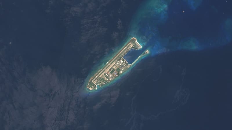 Satellite view of a narrow artificial island with a long runway along its northwestern side, buildings and a harbour basin.