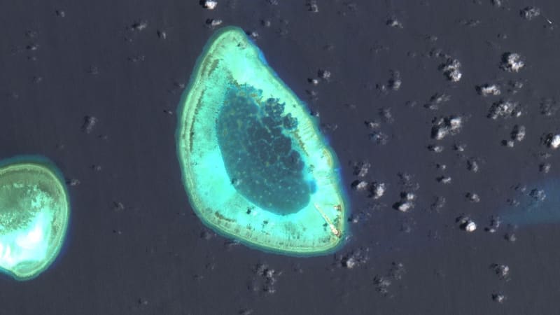 Satellite view of a submerged turquoise coral atoll with a dark lagoon and one small structure on its southeastern rim.
