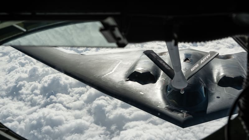 A B-2 Spirit seen from a tanker's boom window above clouds, with the refueling boom connected behind its cockpit