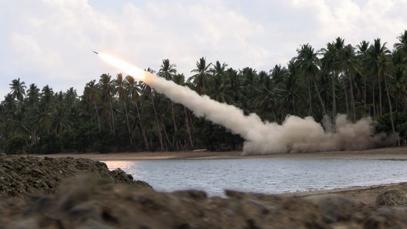 A rocket fired from a HIMARS climbs on a white smoke trail from a palm-lined beach across a lagoon.