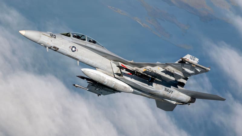 A U.S. Navy F/A-18F Super Hornet banking above clouds and coastline with a dark Blackbeard missile under each wing.