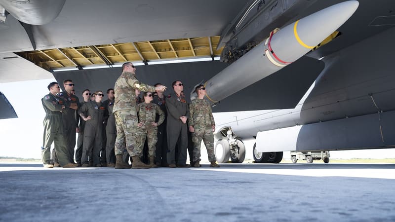Aircrew stand under a B-52 bomber's wing beside a grey-white hypersonic missile with yellow and red bands.