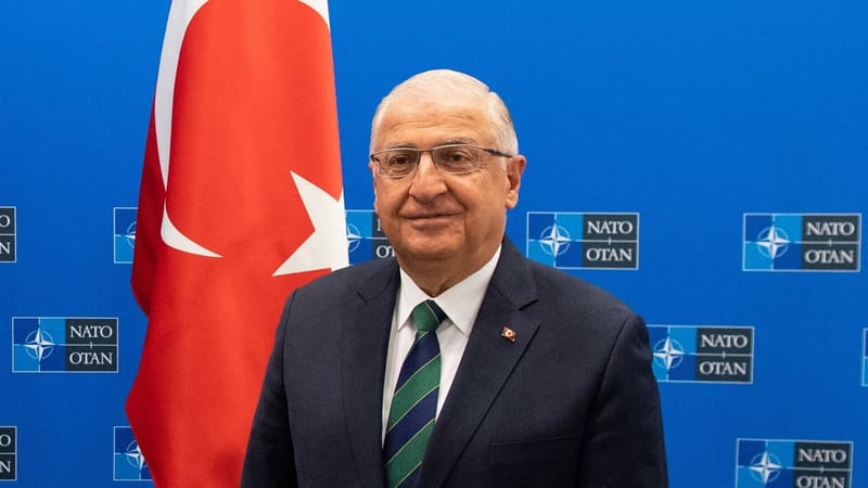 Turkish Defence Minister Yaşar Güler in a dark suit beside a Turkish flag in front of a NATO backdrop.