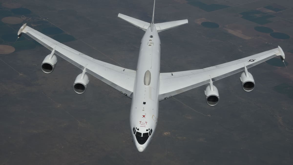 A white U.S. Navy E-6B Mercury four-engine jet seen from above in flight over farmland