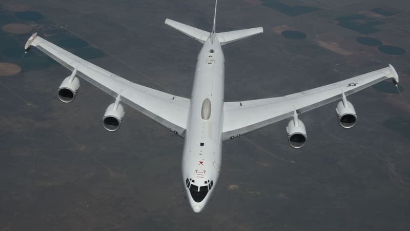 A white U.S. Navy E-6B Mercury four-engine jet seen from above in flight over farmland