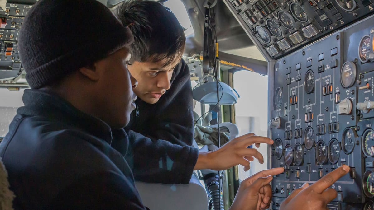 Two U.S. Navy sailors pointing at gauges on the flight engineer panel inside an E-6B cockpit