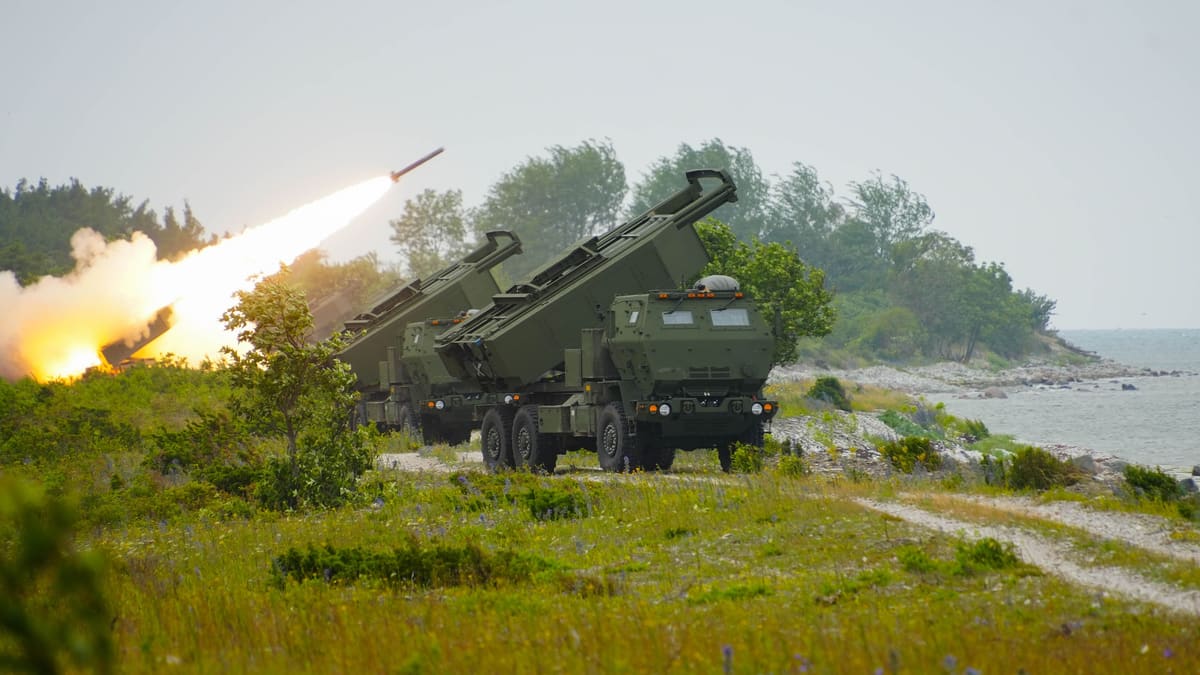 Two Estonian HIMARS rocket launchers with pods raised on a grassy coast as a third fires a rocket behind them