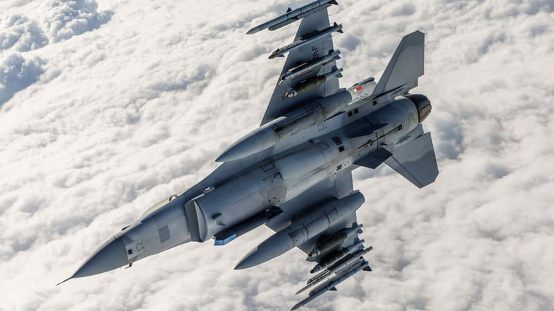 An F-16 Block 70 seen from above over clouds, with missiles, bombs and fuel tanks carried outside the airframe