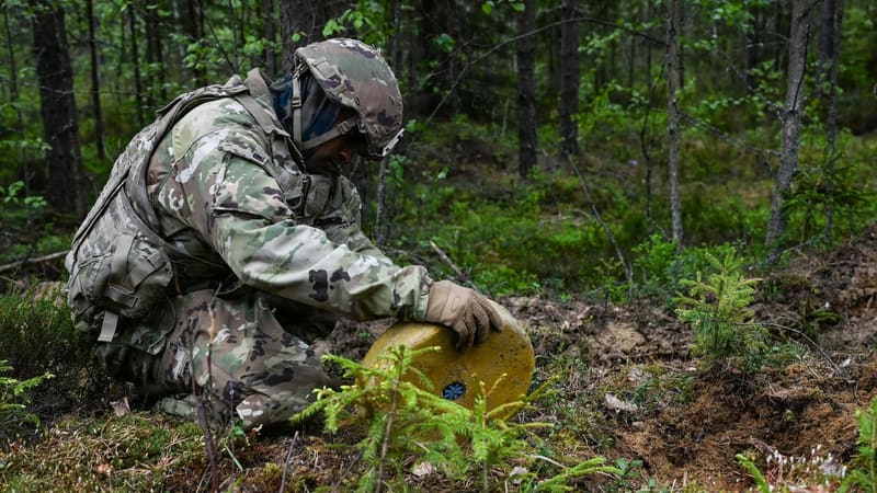 A U.S. soldier in camouflage kneels in a forest, holding a round yellow training mine upright beside freshly dug earth.