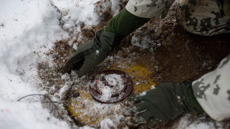 Gloved hands of a Finnish soldier in snow camouflage press a mine into a hole dug in snowy ground.