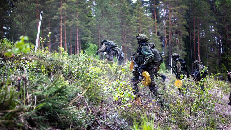 Finnish soldiers in camouflage carry round yellow training mines along a forest path during an exercise.
