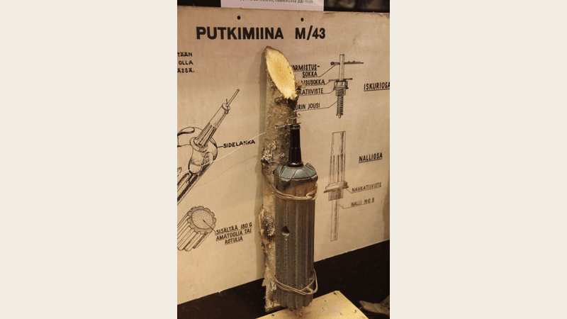 A Finnish m/43 tube mine strapped to a wooden stake stands before a diagram of its parts in a museum display.