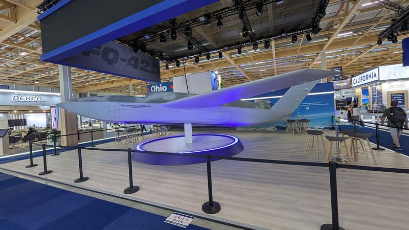Full-scale white and grey General Atomics YFQ-42A drone mock-up on a lit pedestal at an air show stand