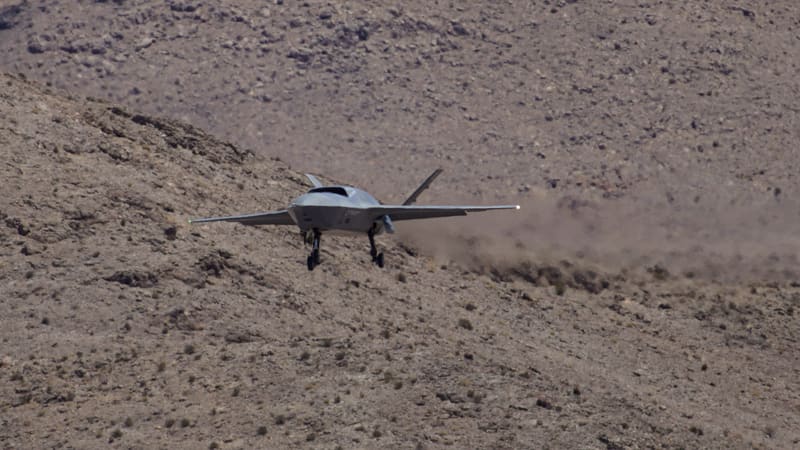 Grey YFQ-42A drone with landing gear down flying low over a brown desert hillside before landing