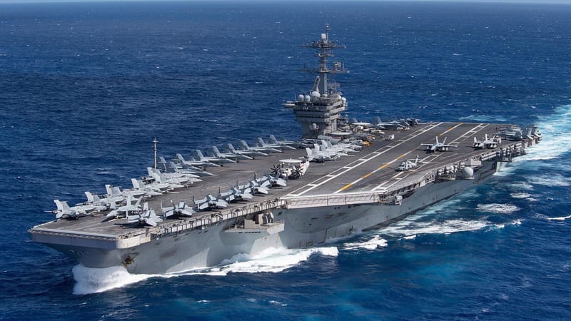 Aerial view of a US Navy aircraft carrier at sea, flight deck lined with jets, white wake at the bow