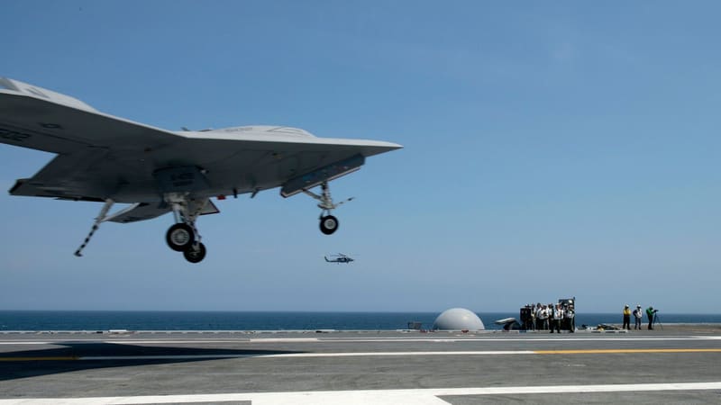 Grey tailless X-47B drone with landing gear down about to touch down on an aircraft carrier deck