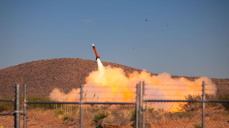 Orange Patriot interceptor climbs out of a cloud of launch smoke over a desert hill, seen through a fence