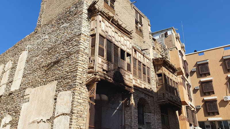 An old stone house with dark wooden latticed balconies beside newer buildings in old Taif.