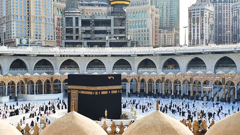 The black-draped Kaaba in the white courtyard of Mecca's Great Mosque, with tall hotel towers rising behind it.