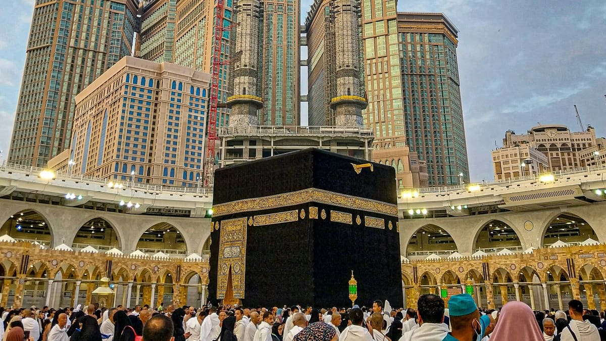 Pilgrims, many in white ihram, crowd around the black-and-gold Kaaba in Mecca, with hotel towers and minarets behind