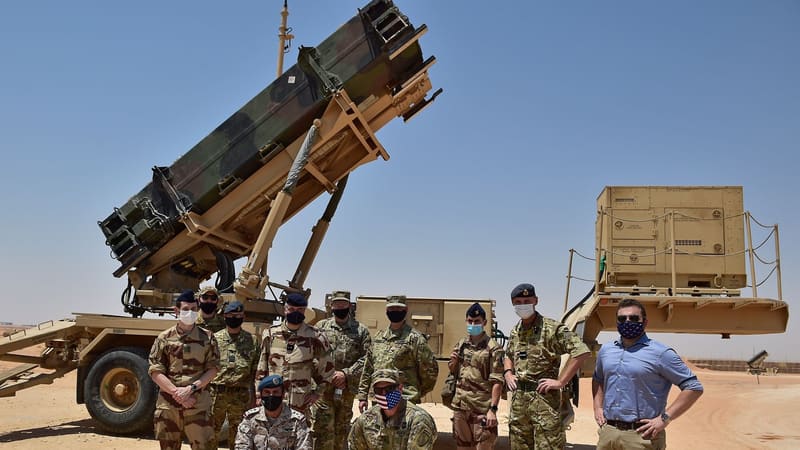 A raised Patriot missile launcher on a desert trailer, with masked military personnel standing and kneeling in front