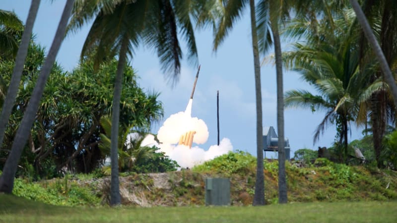 A THAAD interceptor rises from its launcher in white exhaust, framed by palm trees under a blue sky