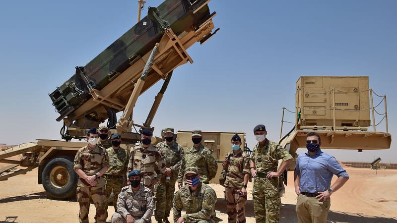 Military personnel and a civilian in face masks pose in front of a raised Patriot missile launcher in the desert