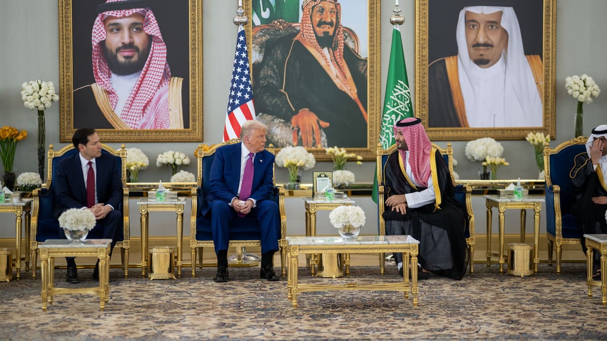 Donald Trump in a blue suit and Mohammed bin Salman in a red-checked headdress talk beneath portraits of Saudi royals