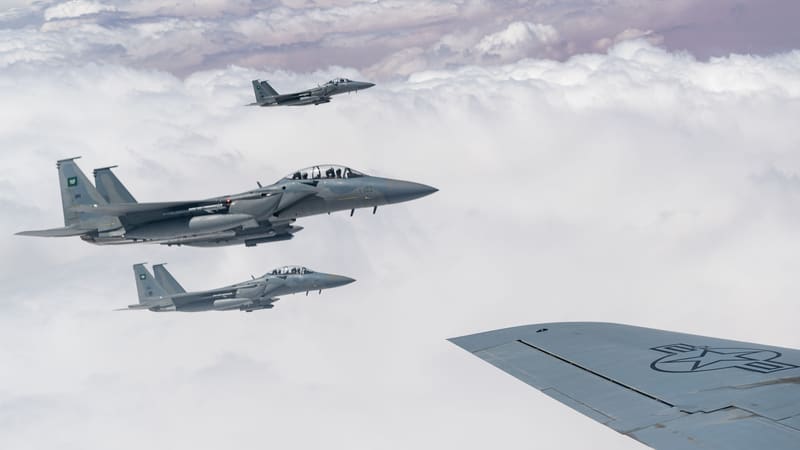 Three Royal Saudi Air Force F-15 fighter jets above clouds, with the wing of a U.S. Air Force aircraft in the foreground