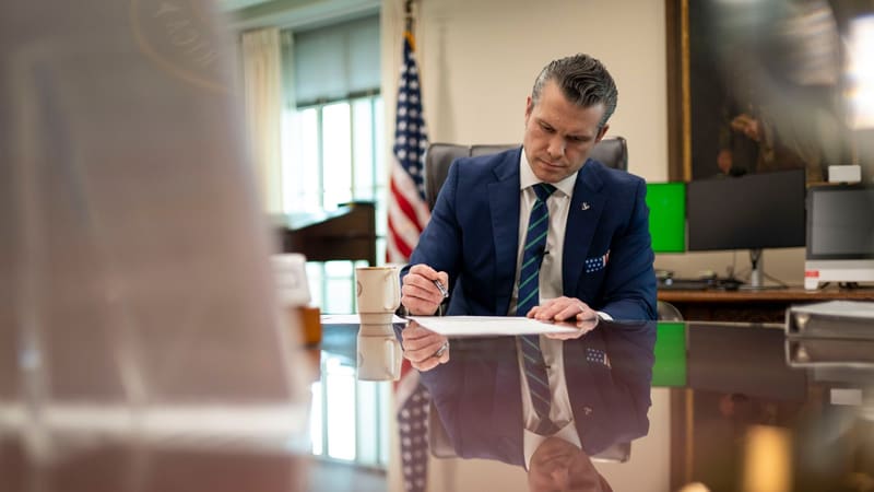 Pete Hegseth signs a document at a polished desk in his Pentagon office with a U.S. flag behind him