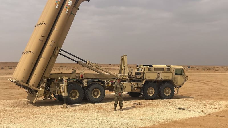 A soldier stands in front of a tan THAAD launcher truck with its interceptor canisters raised in the desert