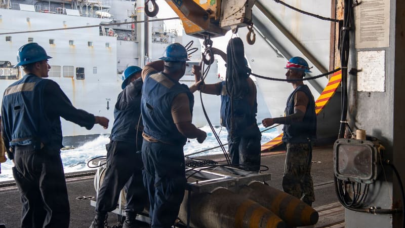 Navy sailors in helmets guide yellow-banded bombs on a hoist inside a carrier hangar bay beside a supply ship