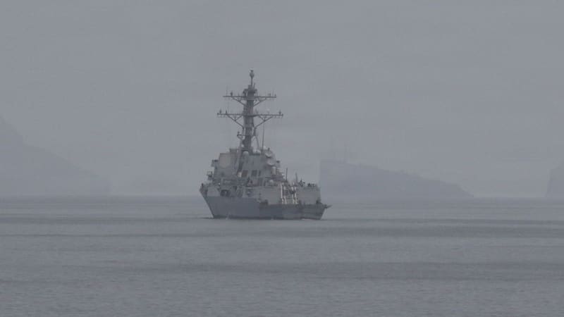 US Navy destroyer on calm grey water in thick haze, with rocky islands faint behind it