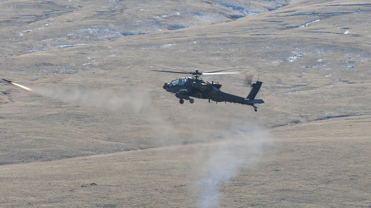 A U.S. Army AH-64E Apache flying low over brown hills as a just-fired AGM-114 Hellfire missile streaks away at left