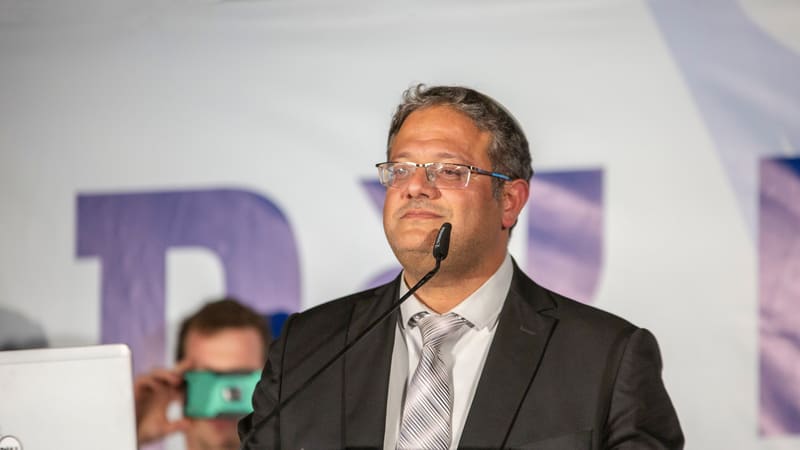 Man in glasses and a dark suit at a podium with a microphone, in front of a purple and white banner