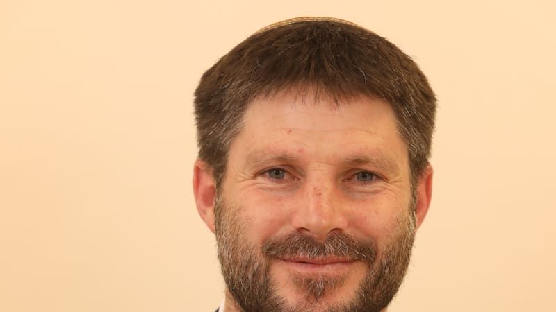 Bearded man in a navy jacket and white shirt wearing a kippah against a cream background