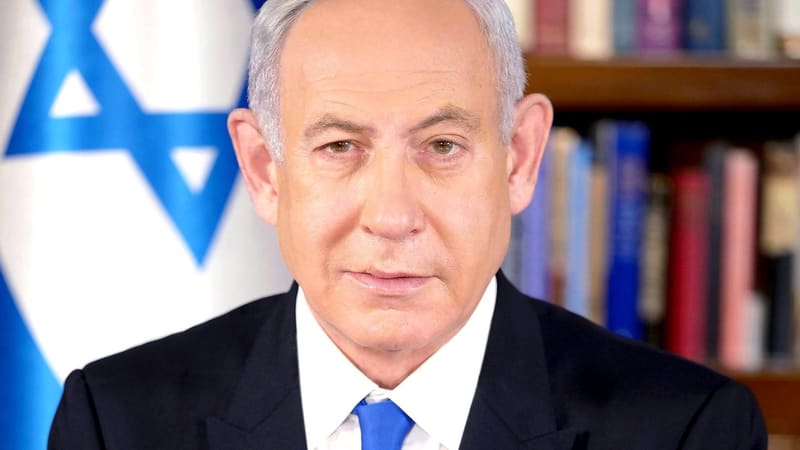 Benjamin Netanyahu in a dark suit and blue tie in front of an Israeli flag and bookshelves