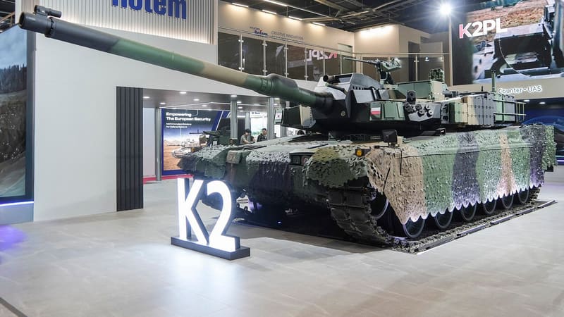 A camouflaged K2PL tank with a Polish flag on its turret, on the Hyundai Rotem stand behind an illuminated K2 sign