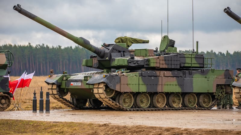 A camouflaged Polish Army K2 Black Panther tank with a Polish flag on an antenna, parked on a muddy training range