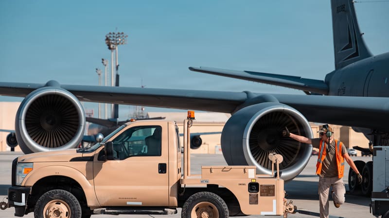 A maintainer in an orange vest beside a KC-135's engine and a tan service truck on a desert airfield.